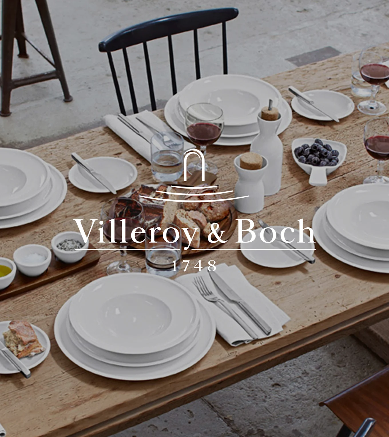 A rustic wooden table set with Villeroy & Boch white dinnerware, wine, bread, blueberries, salt, pepper, and olive oil for a meal.