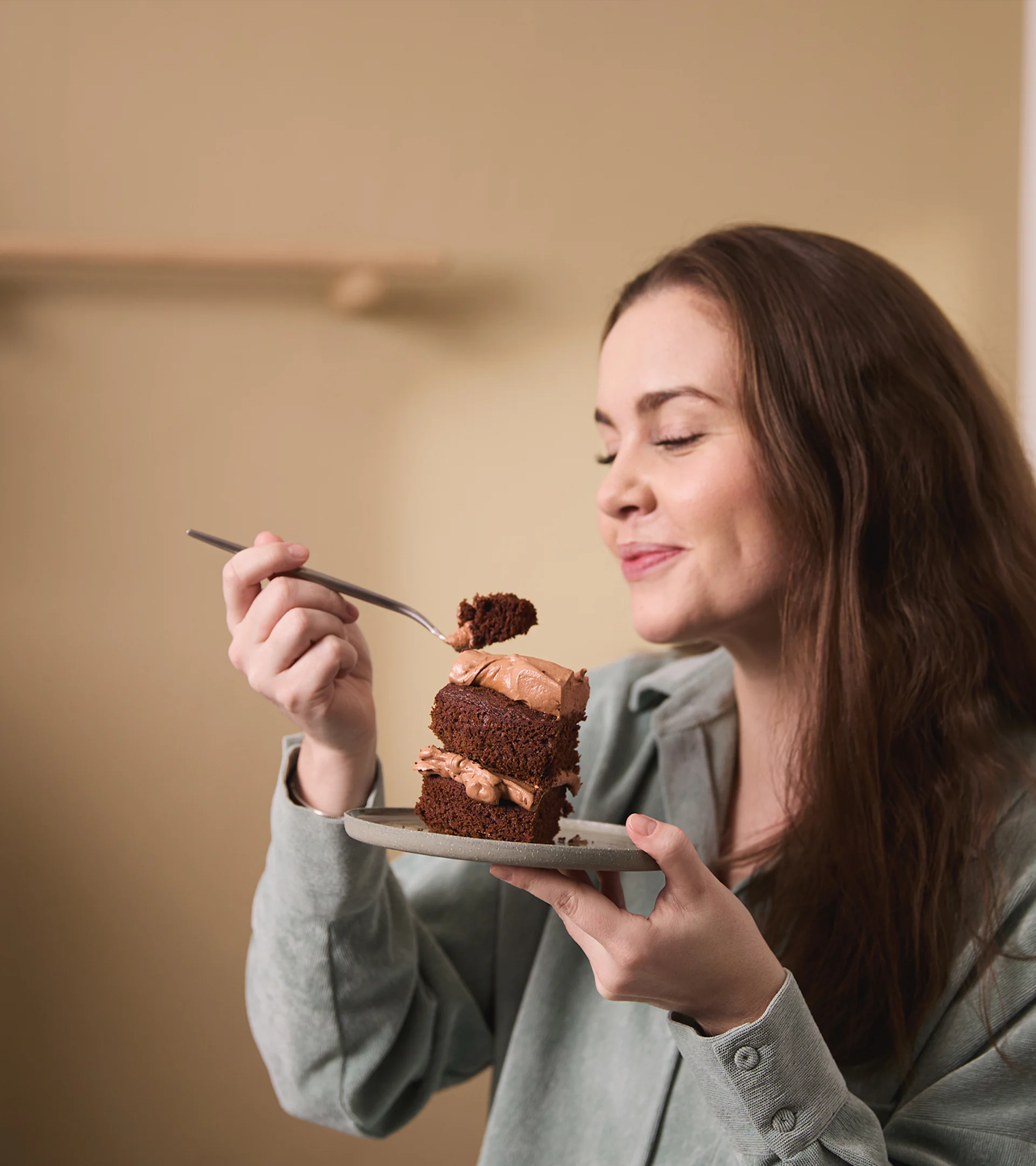 Woman with long brown hair, eyes closed, enjoying a bite of chocolate cake from a small plate.
