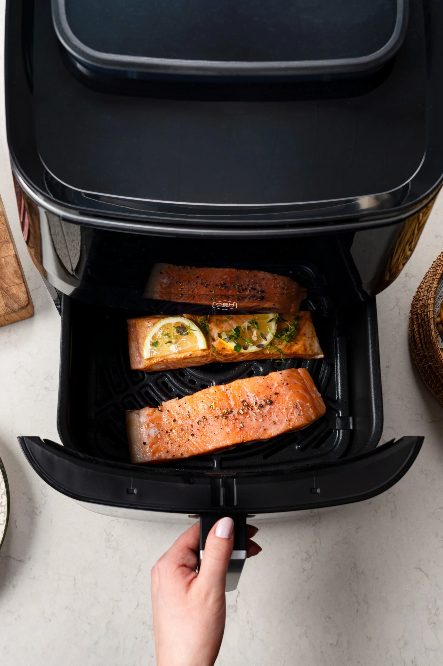A hand pulls out the drawer of a black air fryer, revealing three pieces of seasoned salmon, one with lemon slices and herbs.