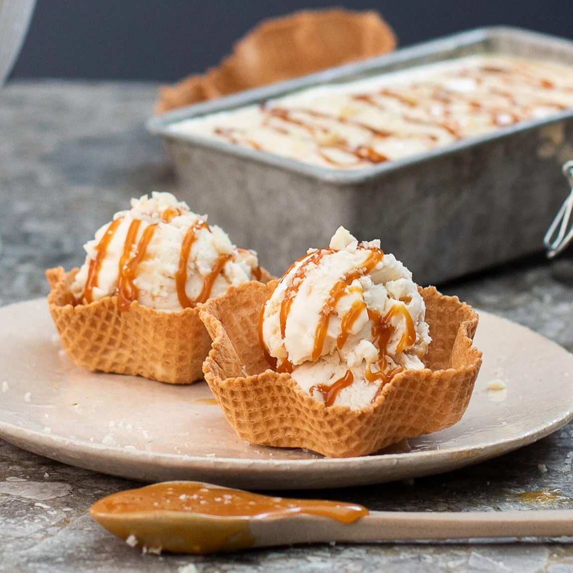 Two scoops of ice cream with caramel sauce in waffle bowls, with a pan of ice cream in the background.