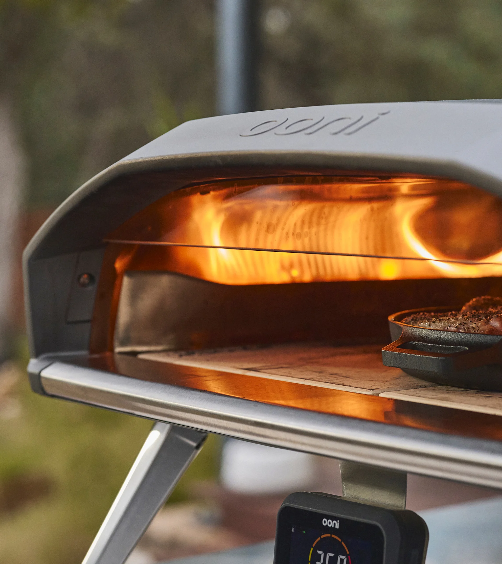 A close-up of a pizza oven with roaring flames inside, cooking food in a cast iron pan, with a digital thermometer below.