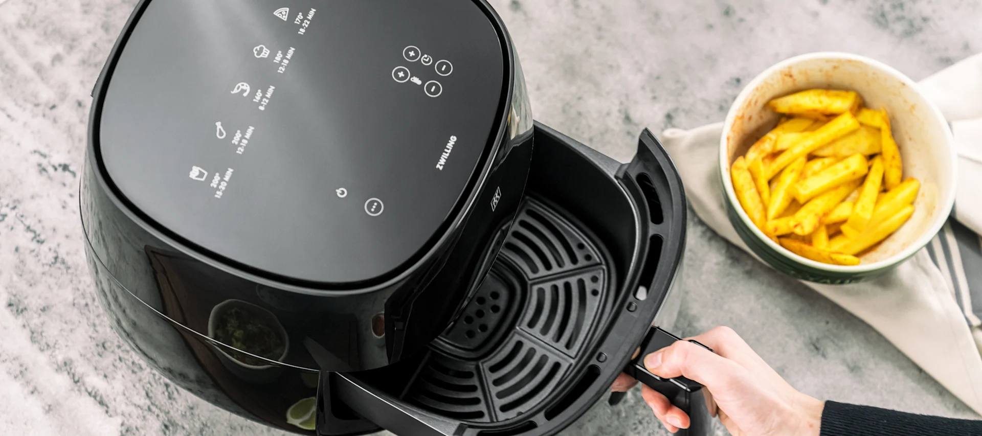 Overhead view of a hand pulling out the basket of a black air fryer, next to a bowl of french fries.