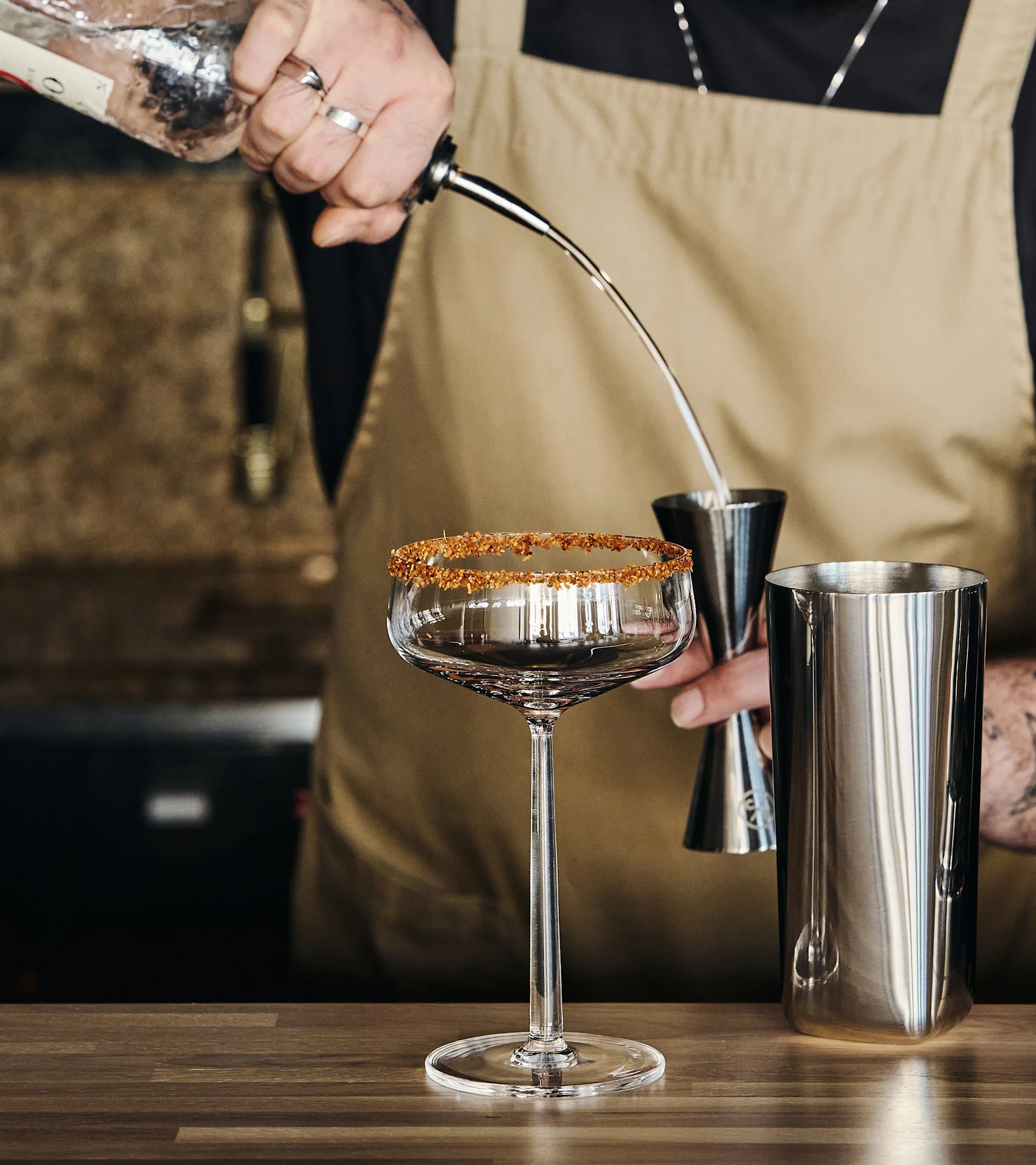A bartender pouring a drink from a bottle into a jigger, with a sugar-rimmed cocktail glass and a shaker on a wooden counter.