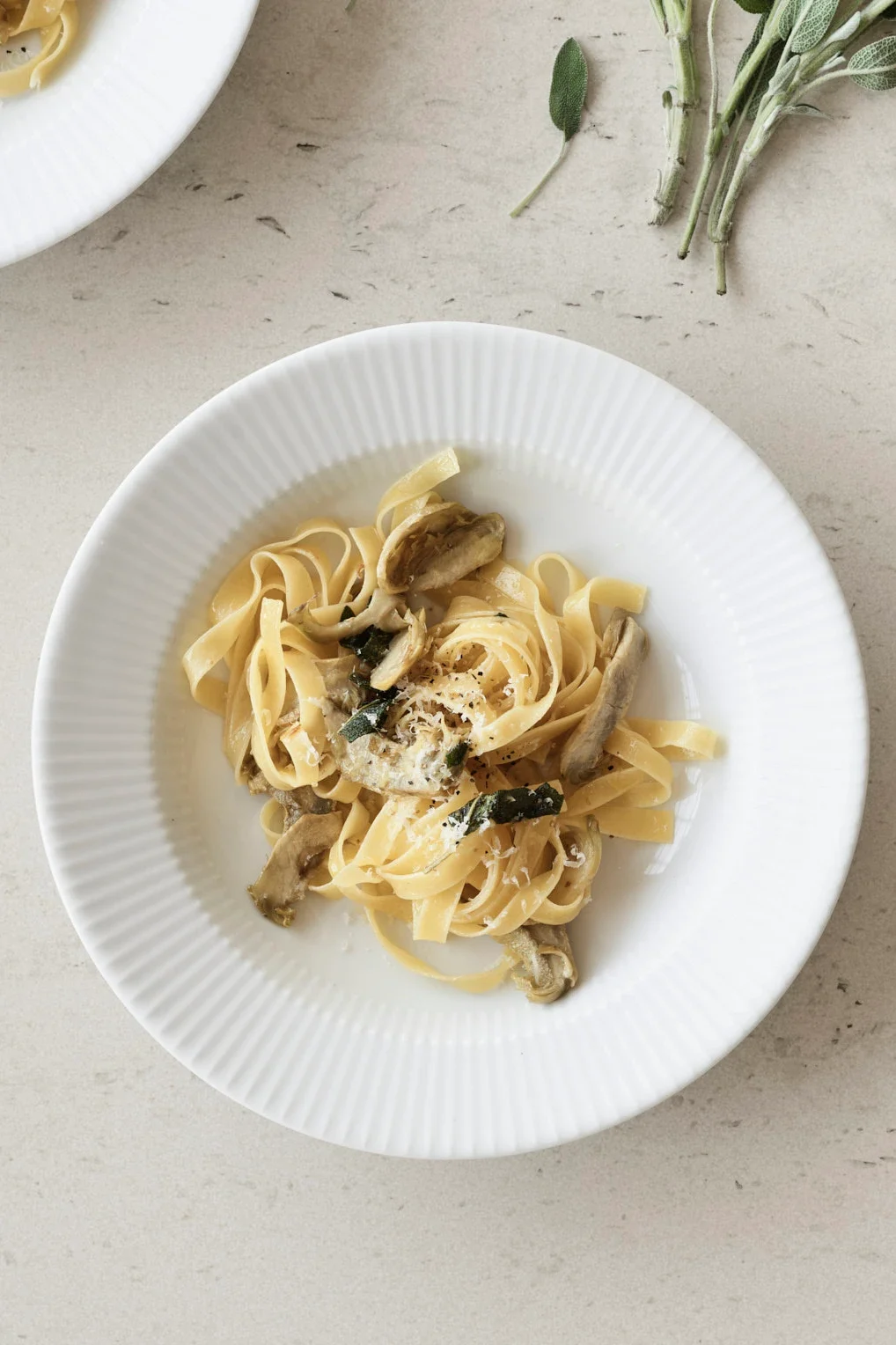 Pasta with artichokes, sage, and grated cheese on a white plate, with sage sprigs on a light counter.