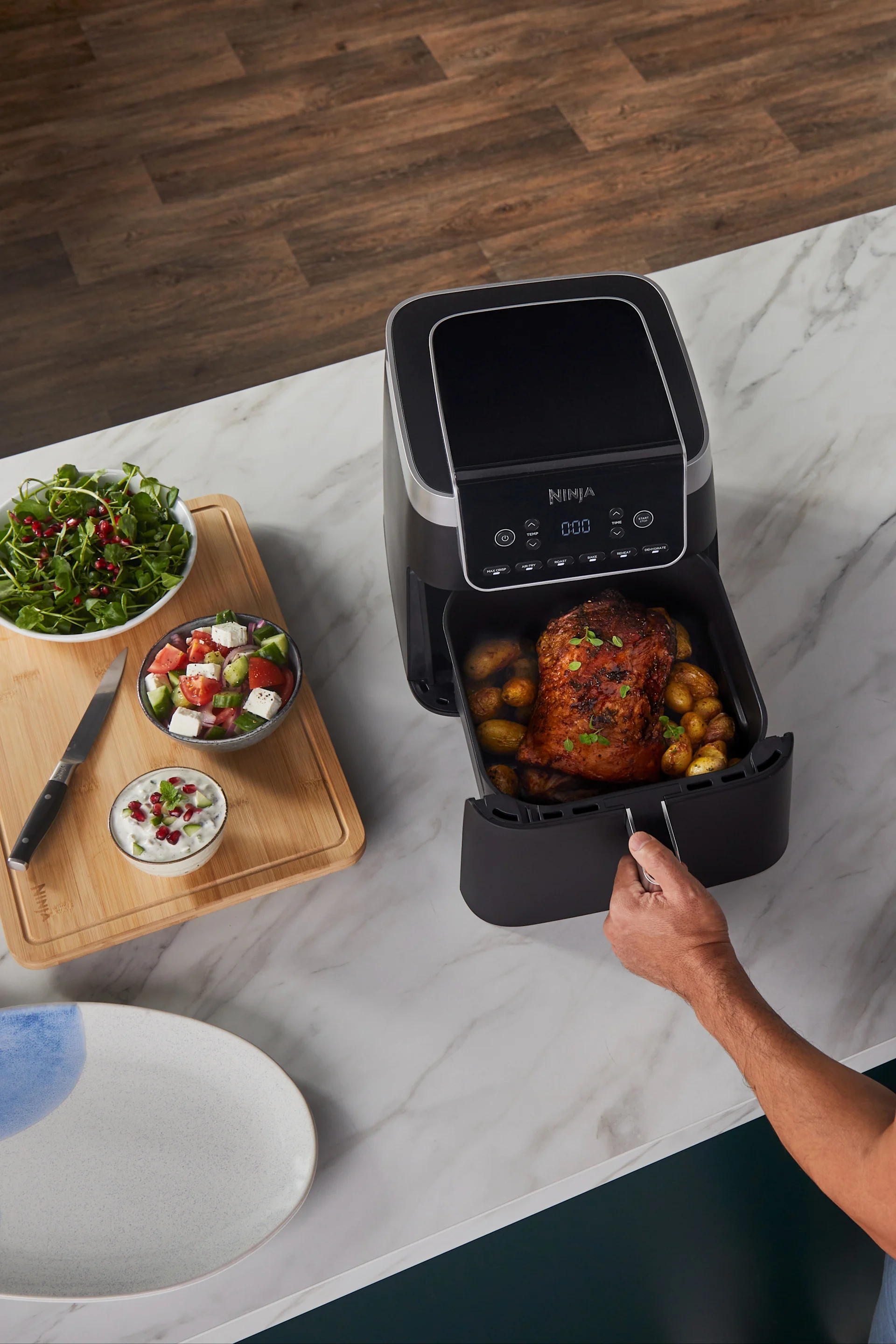 Overhead view of a person pulling out a black air fryer drawer with a roasted chicken and potatoes, next to salads and a dip.
