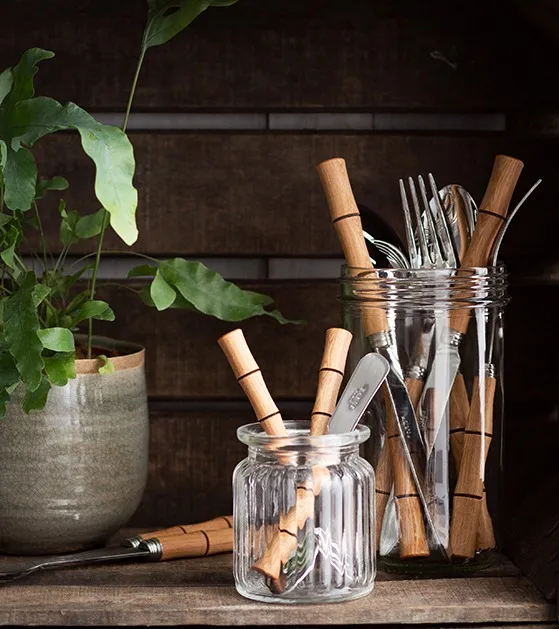 Cutlery with wooden handles in glass jars, next to a potted fern on a dark wooden shelf.