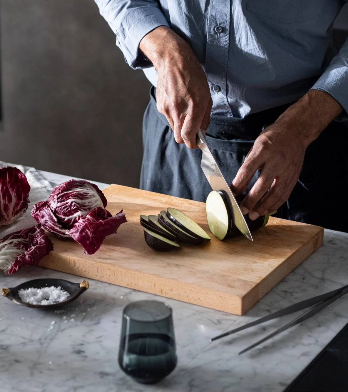 Close-up of a person slicing eggplant on a wooden cutting board with radicchio and salt nearby.