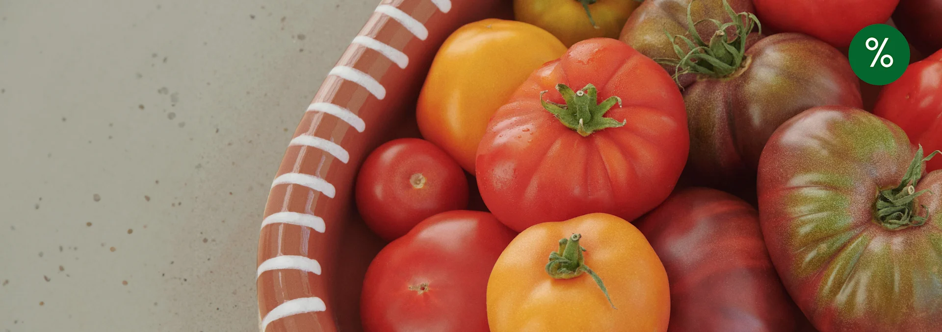 Various ripe red, yellow, and heirloom tomatoes in a terracotta bowl with a green percentage sign.