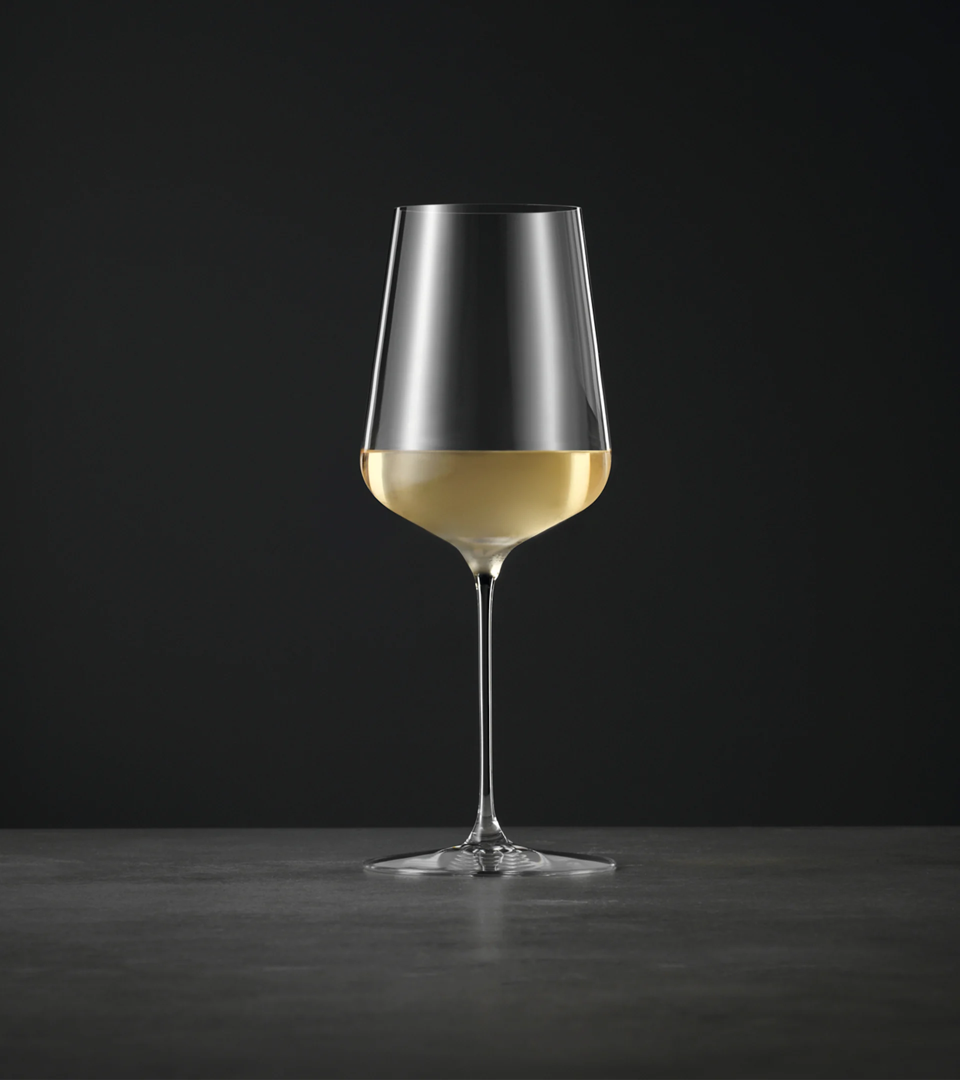 A tall, elegant wine glass with a long stem, half-filled with light white wine, against a dark background.