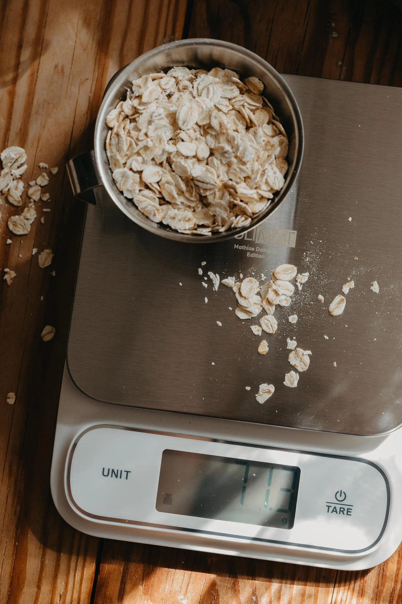 Overhead view of rolled oats in a metal bowl on a digital kitchen scale showing 113g, with some oats spilled on a wooden table.