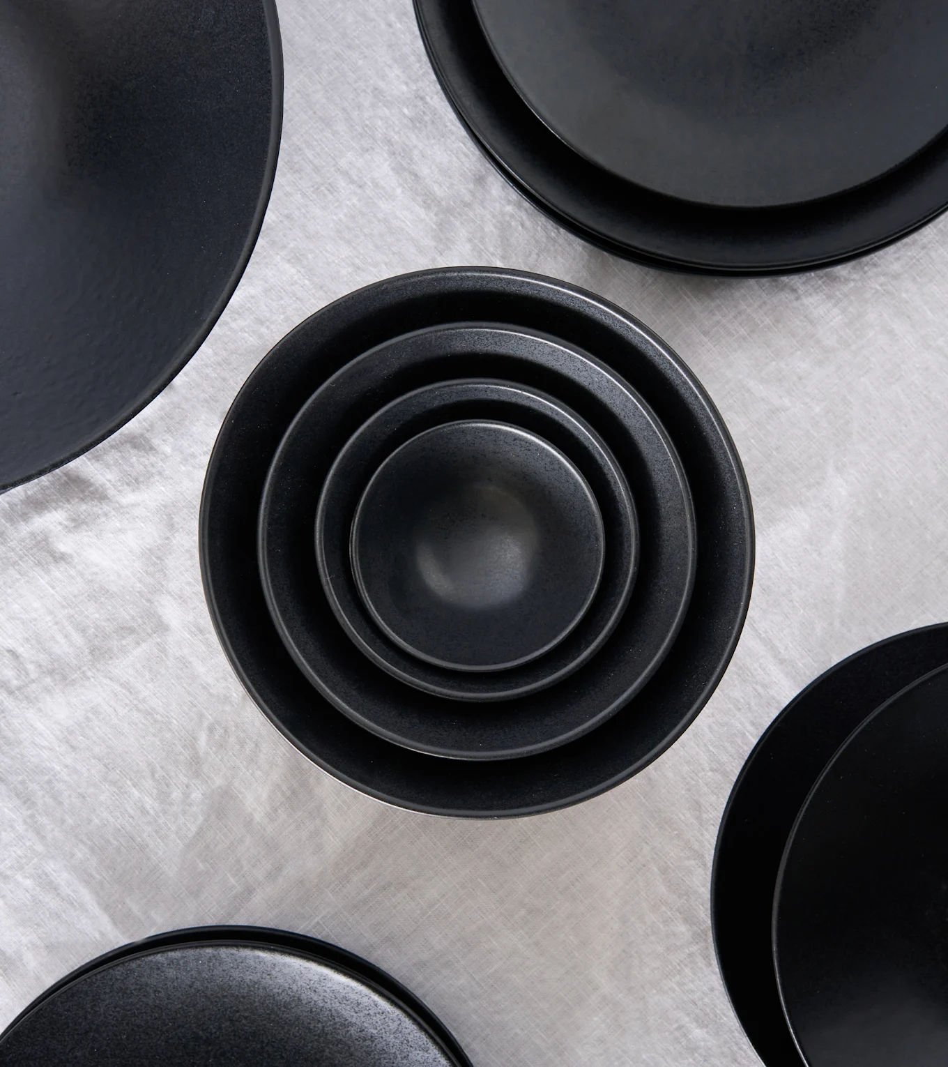Top-down view of black matte ceramic bowls nested in each other, surrounded by other black plates on a light gray fabric.