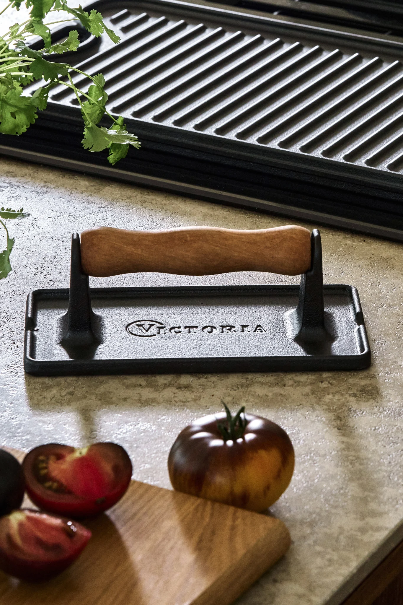 A cast iron meat press placed on a kitchen side next to tomatoes and a griddle cooking plate. 