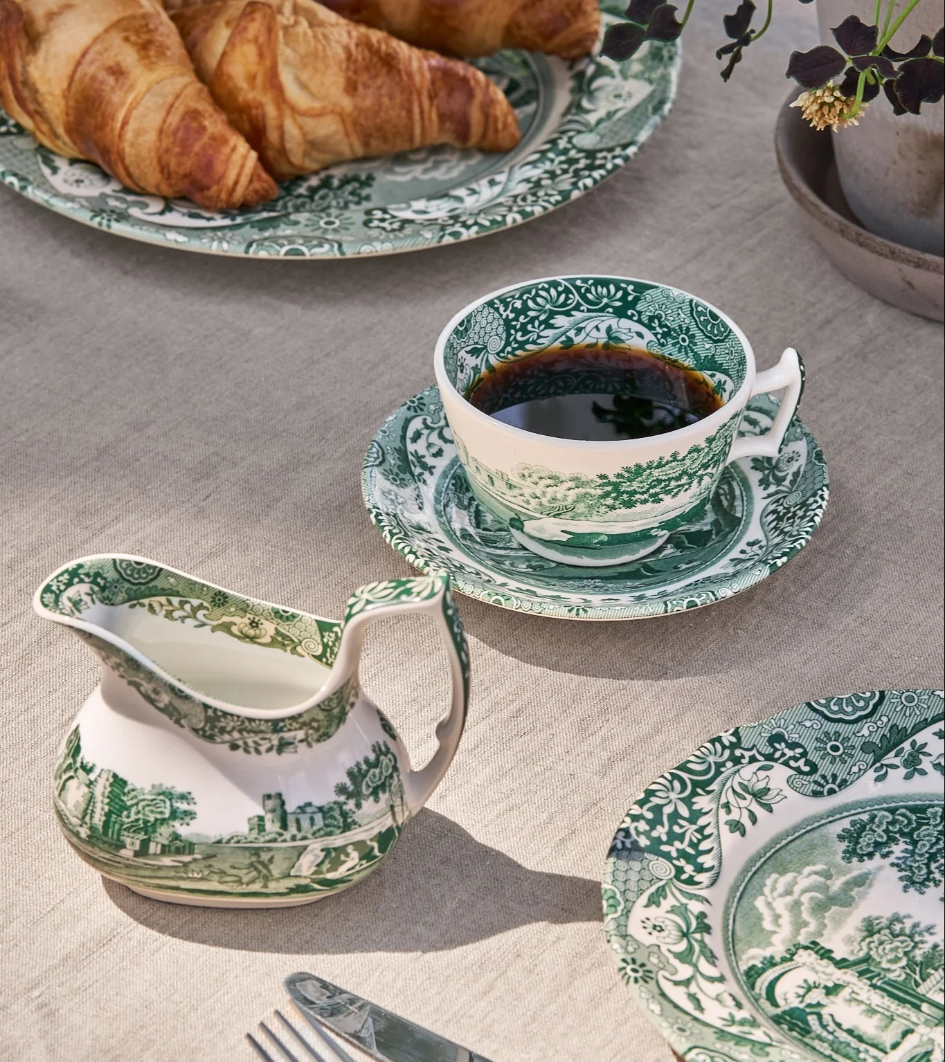Breakfast table with green and white patterned porcelain cup with coffee, creamer, and plates with croissants.