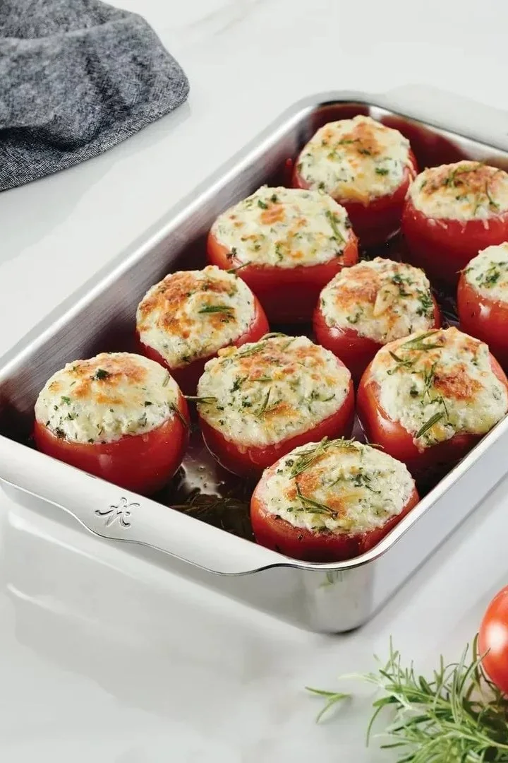 Nine baked red tomatoes stuffed with a creamy, herbed filling in a rectangular stainless steel baking dish.