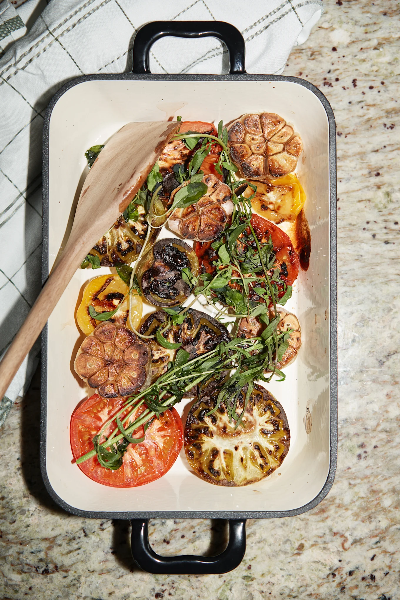 Grilled tomatoes, garlic, and fresh greens in a white baking dish with a wooden spoon, on a granite countertop.