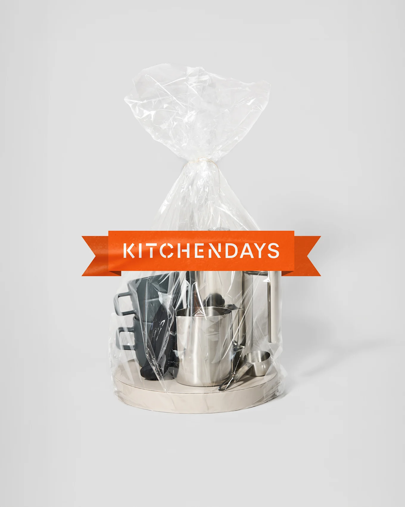 A transparent gift bag tied with string, containing a silver coffee maker, a silver frothing pitcher, and black mugs on a light tray, with an orange "KITCHENDAYS" banner.