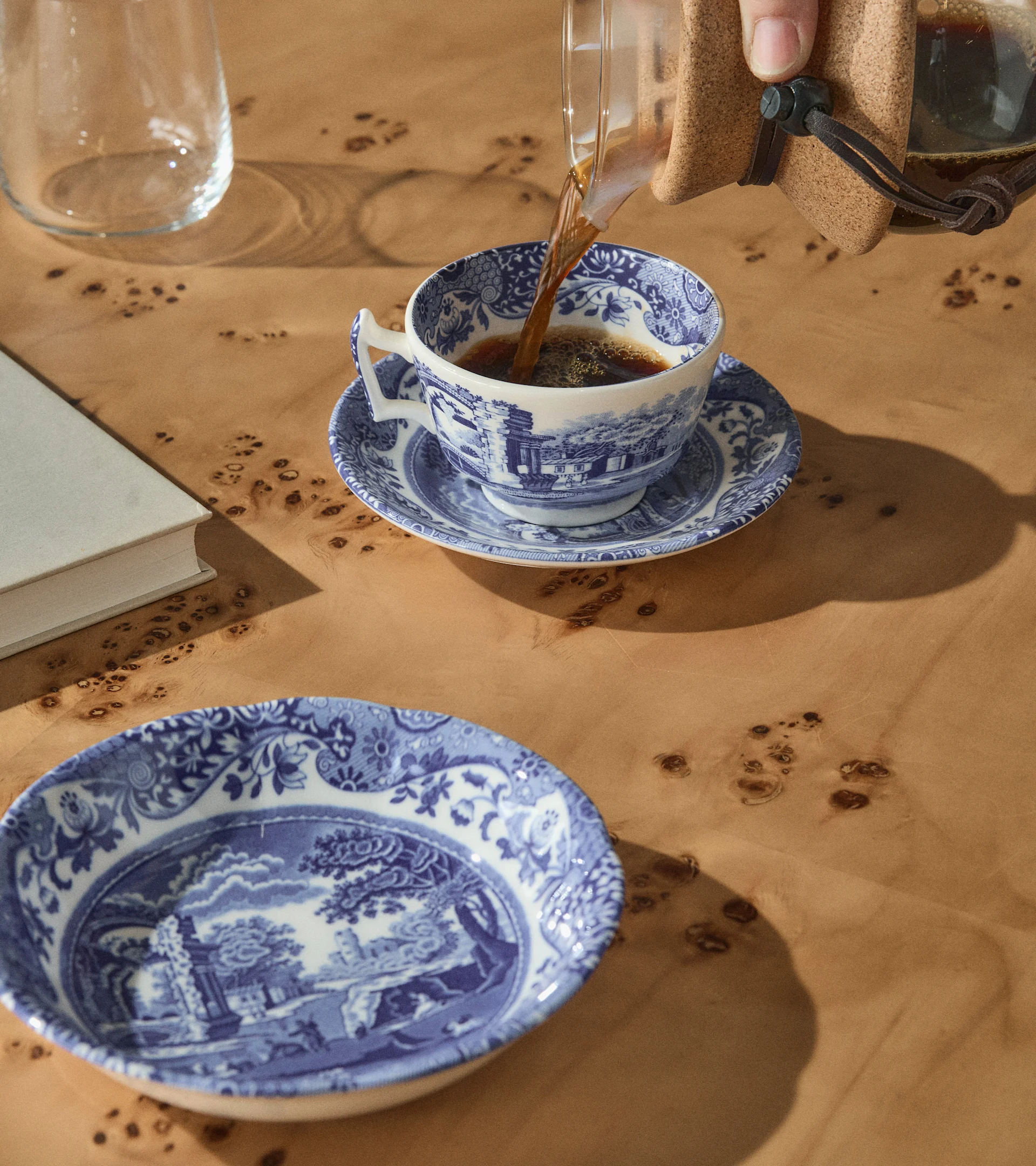 A hand pours coffee into a blue and white patterned teacup on a wooden table with a matching bowl, a book, and a glass.