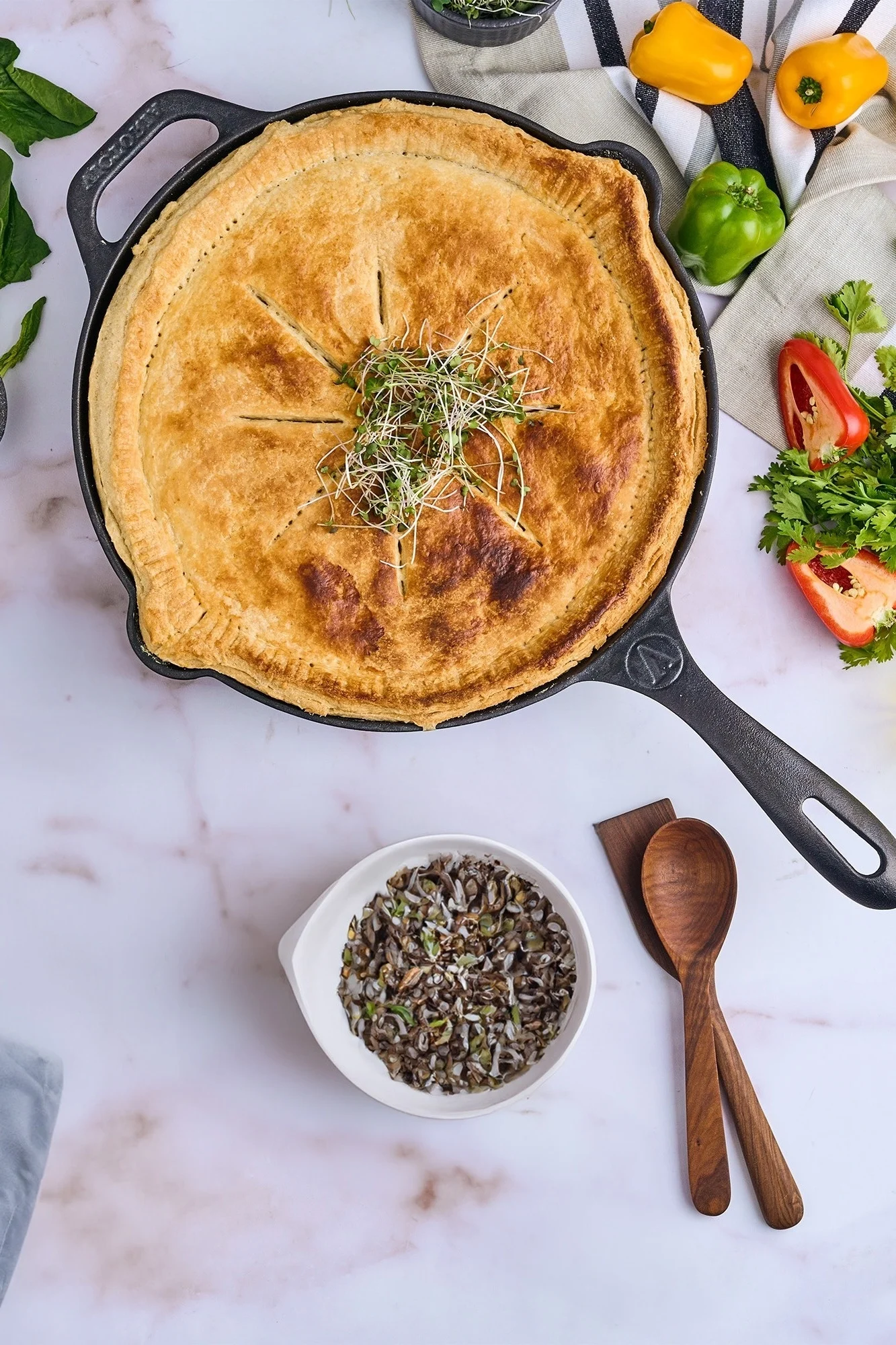 Golden savory pie in cast iron skillet, surrounded by colorful peppers, herbs, spices, and a bowl of wild rice on marble.