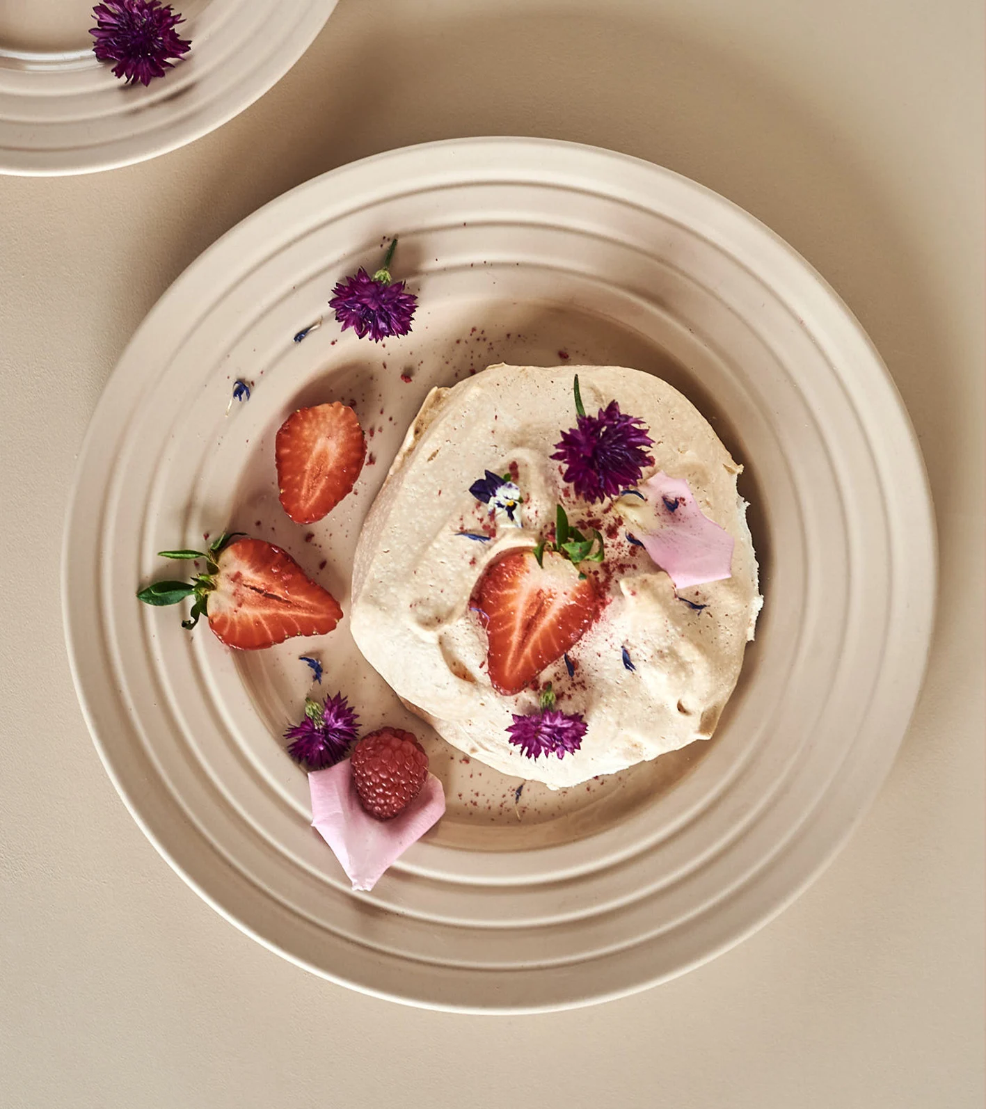 A creamy dessert with strawberries, a raspberry, and purple flowers on a beige ribbed plate.