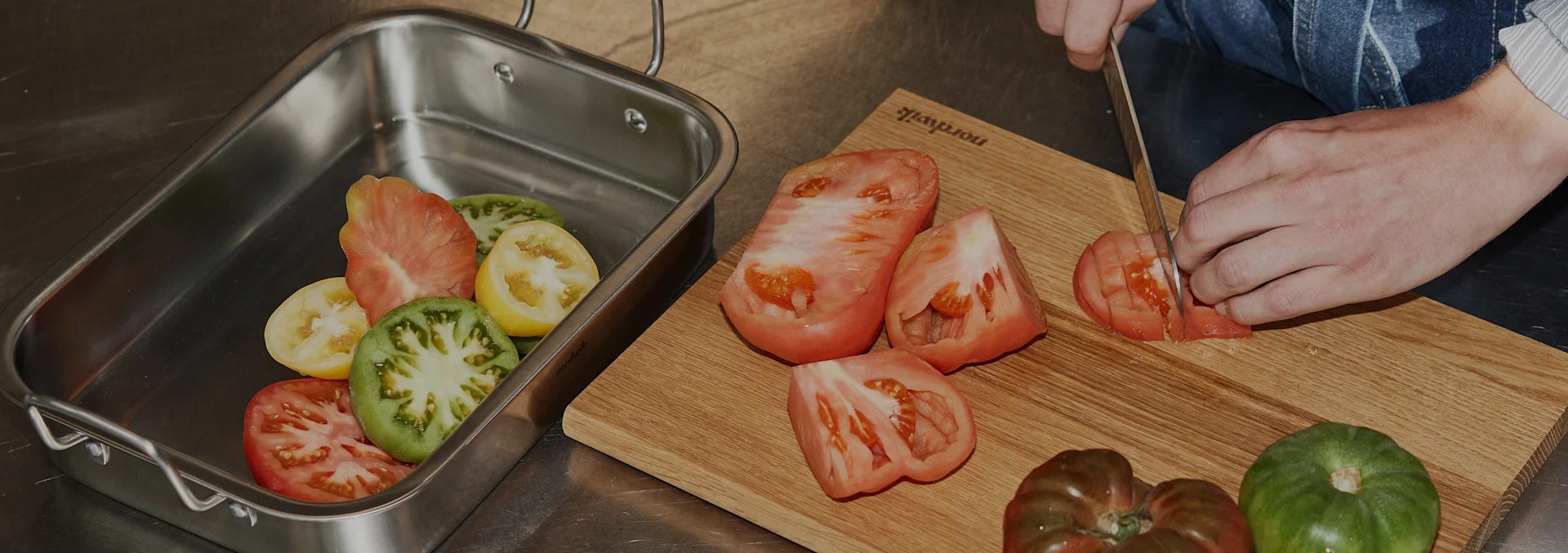 Hands slicing heirloom tomatoes on a wooden cutting board with more tomatoes in a steel roasting pan.