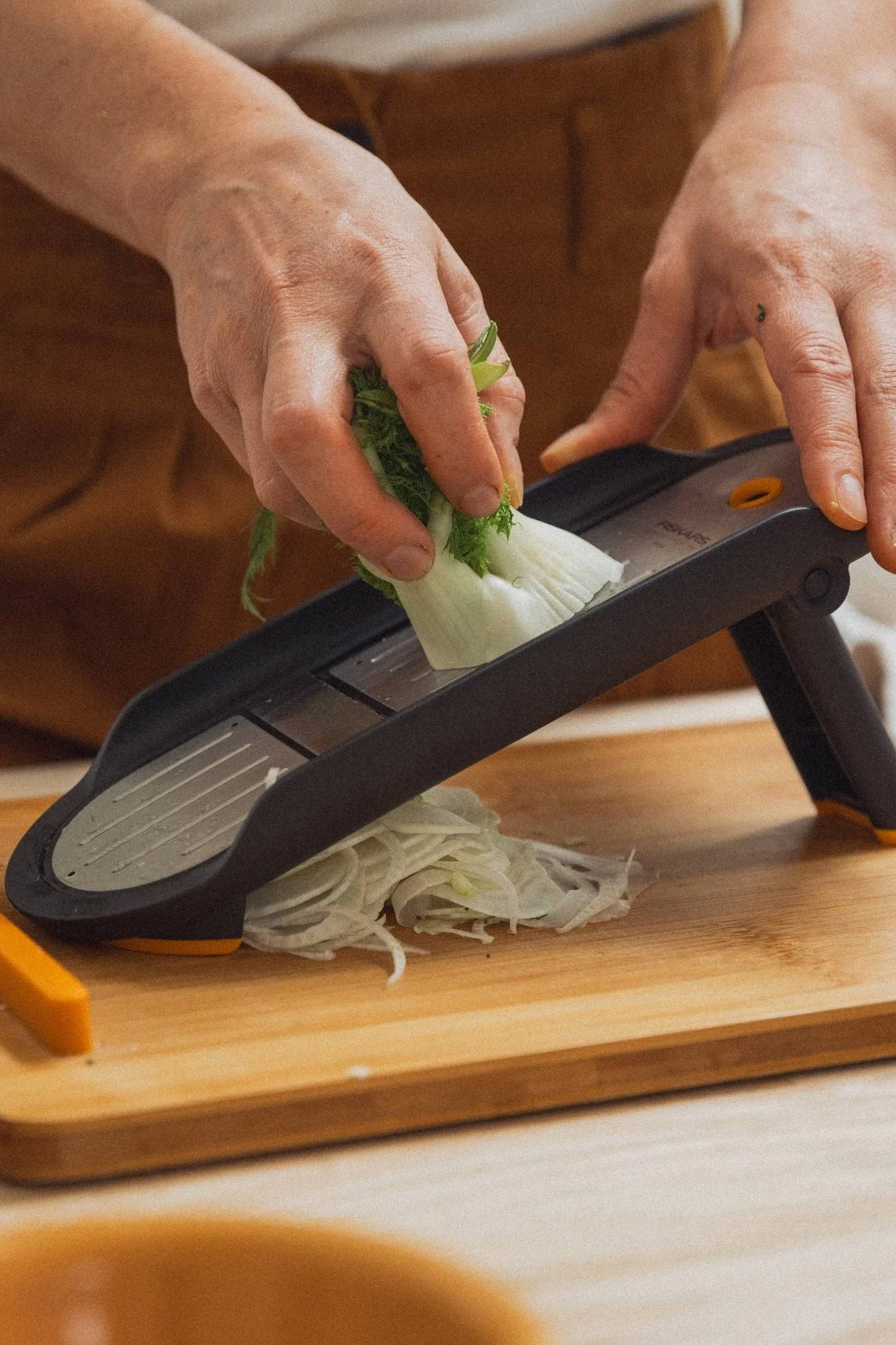 Hands slicing fennel with a grey mandoline slicer on a wooden cutting board.
