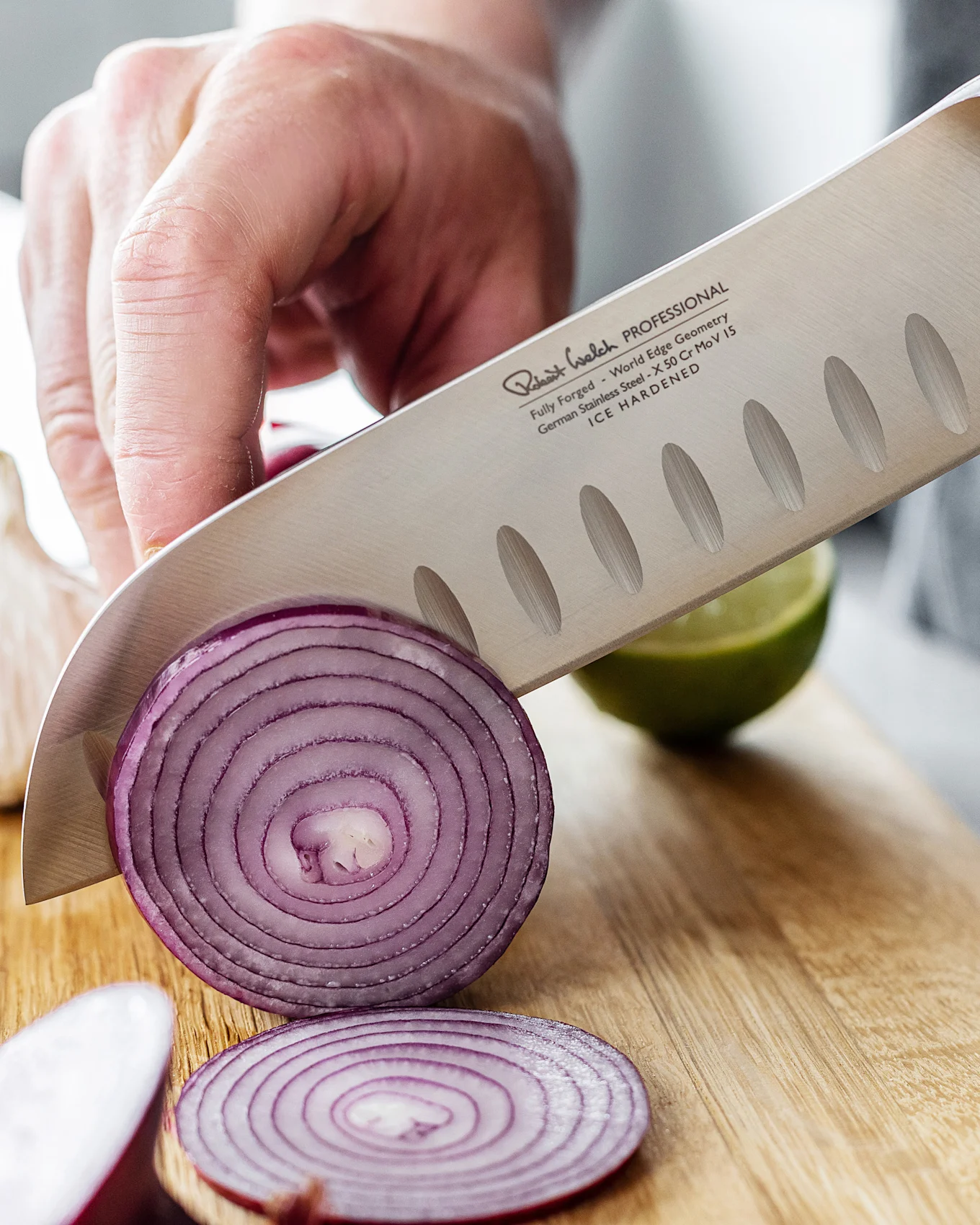 A chef's knife with a textured blade slicing a red onion on a wooden board, with a lime in the background.