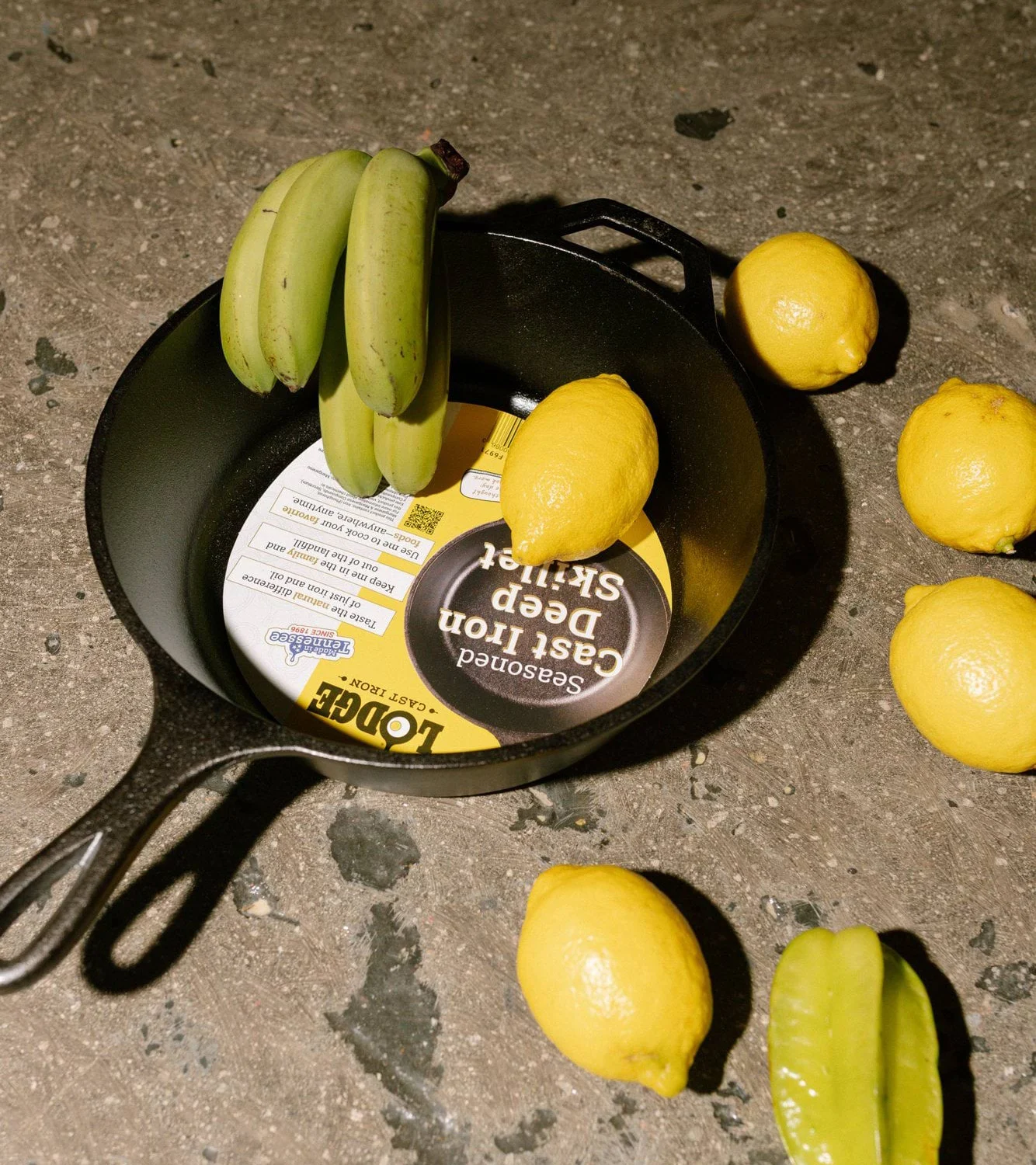 Cast iron skillet with a label inside, unripe bananas, lemons, and a star fruit on a dark surface.
