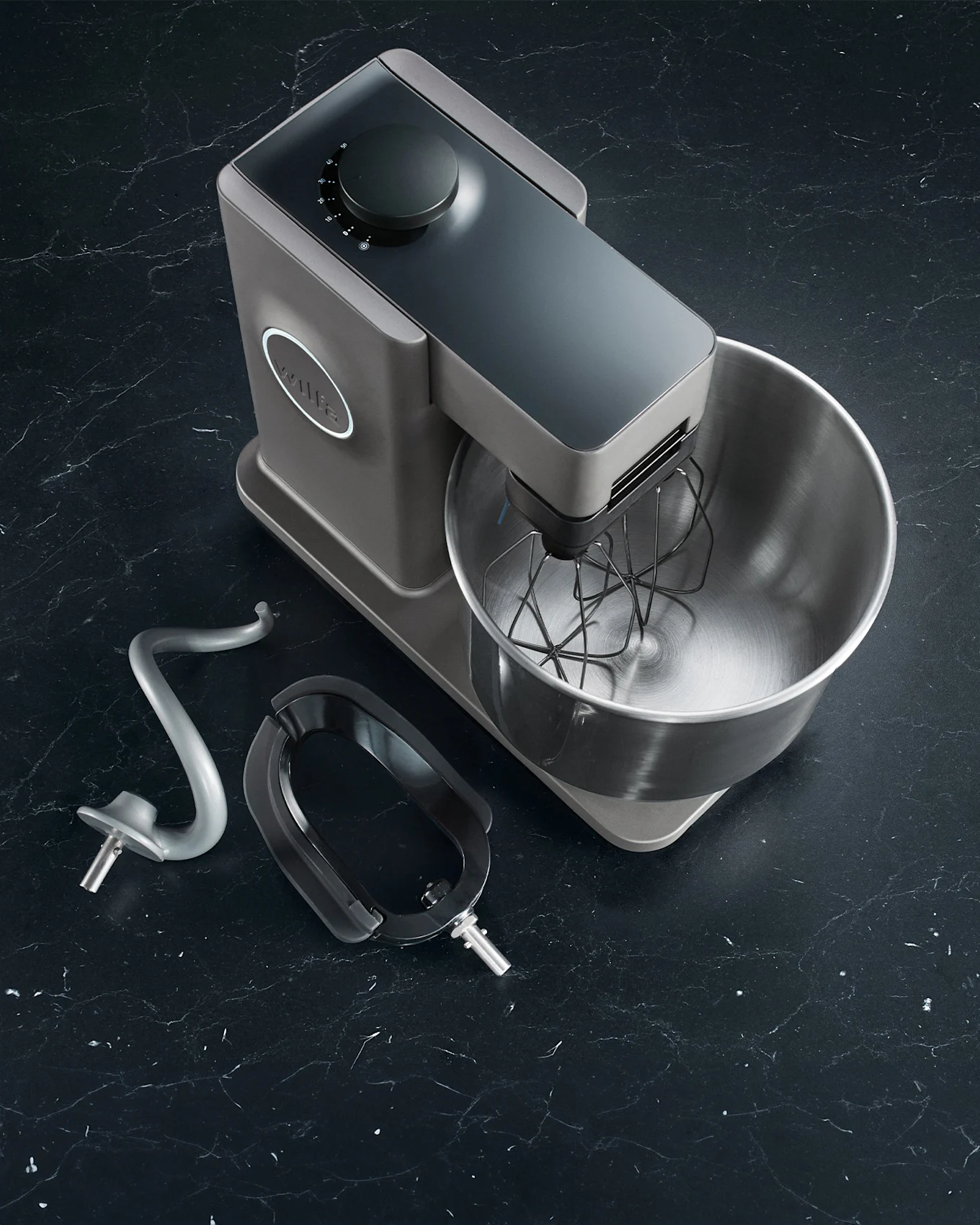 Grey stand mixer with bowl and whisk, plus dough hook and paddle beater, on a dark marble surface.