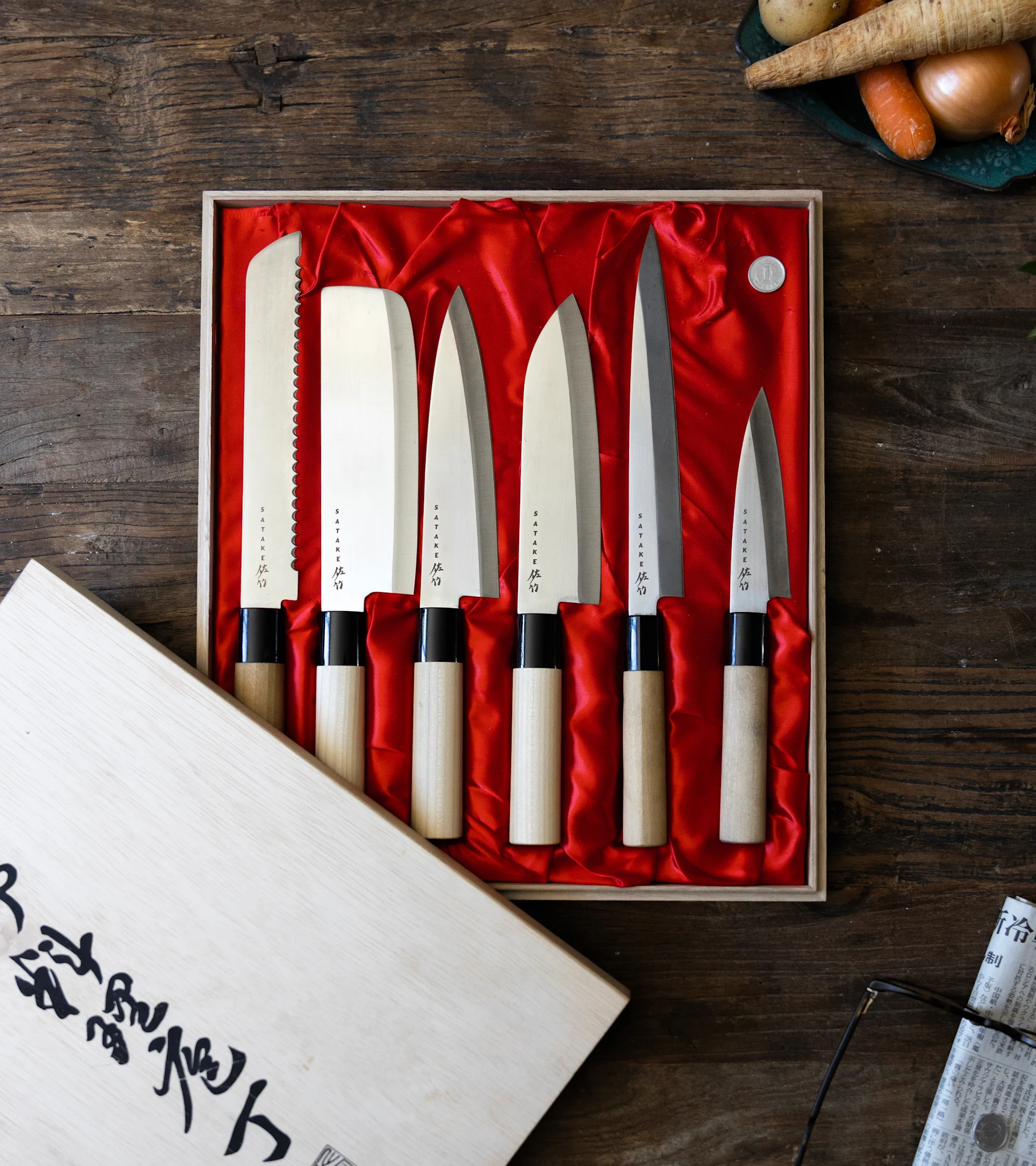 Six Japanese knives with wooden handles in a red-lined box on a wooden table, with vegetables and newspaper in the background.