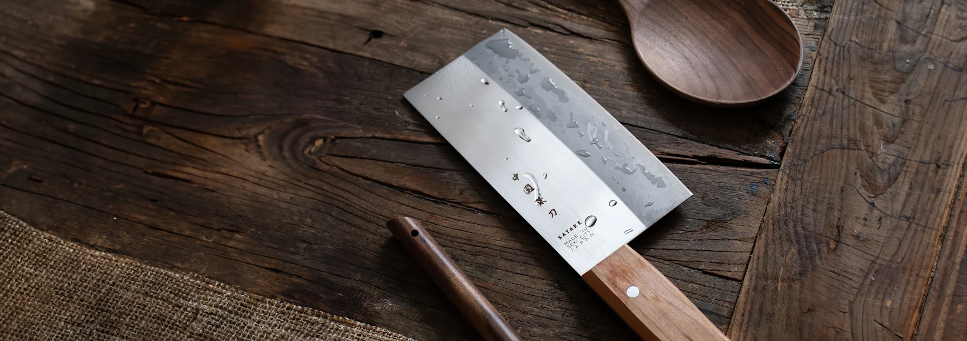 A cleaver-style chef's knife with water droplets on its blade and a light wooden handle, lying on a rustic wooden surface with a wooden spoon.
