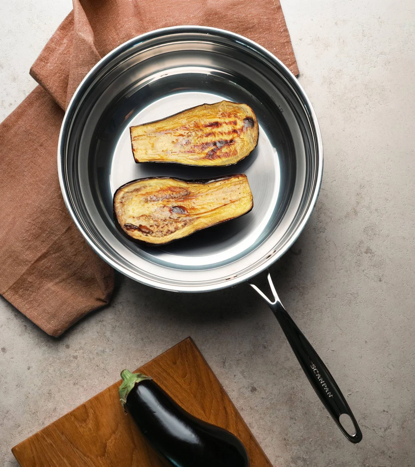 Two grilled eggplant halves in a stainless steel frying pan, with a raw eggplant on a cutting board nearby.