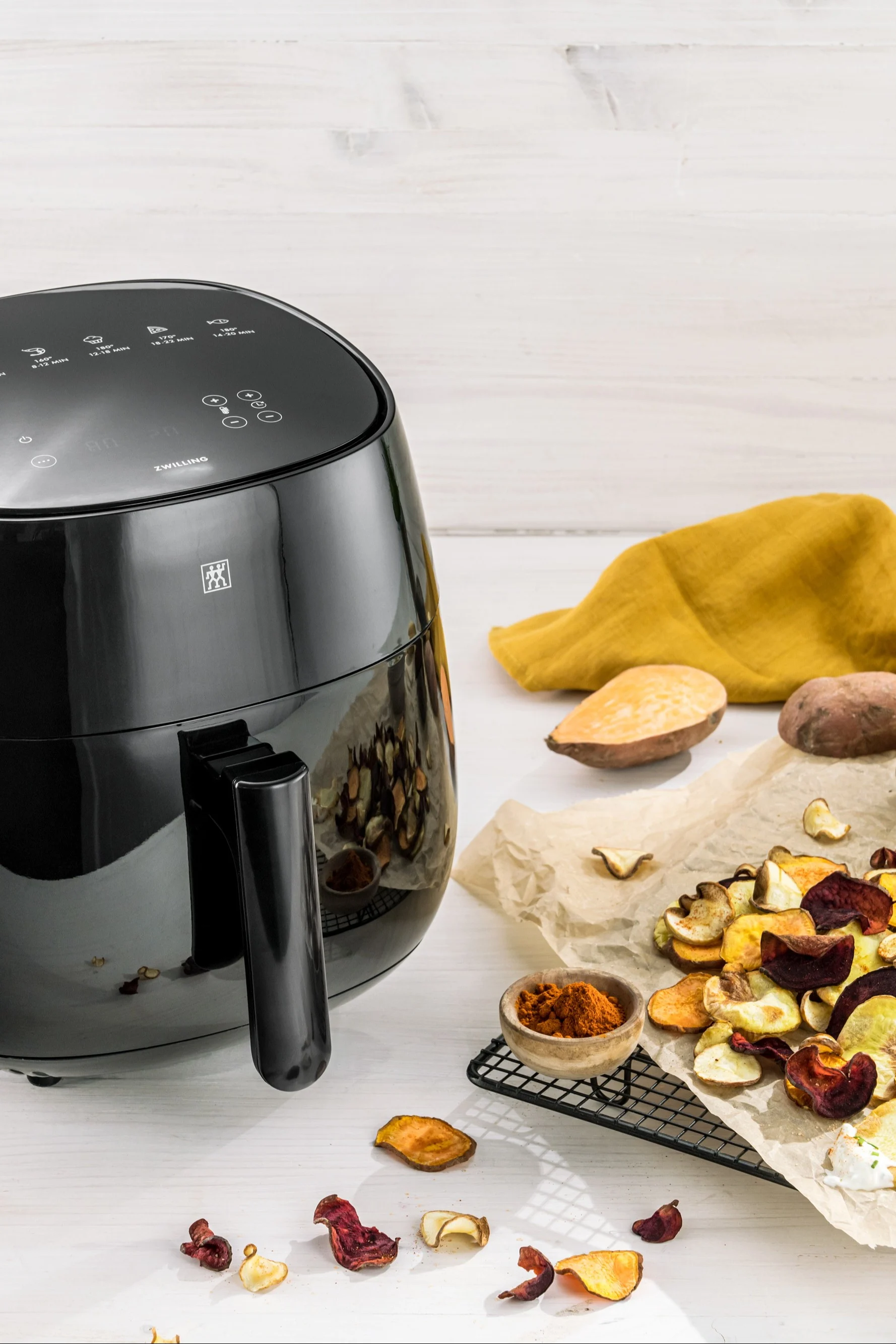 Black air fryer next to a pile of colorful homemade root vegetable chips and spices on a white wooden table.