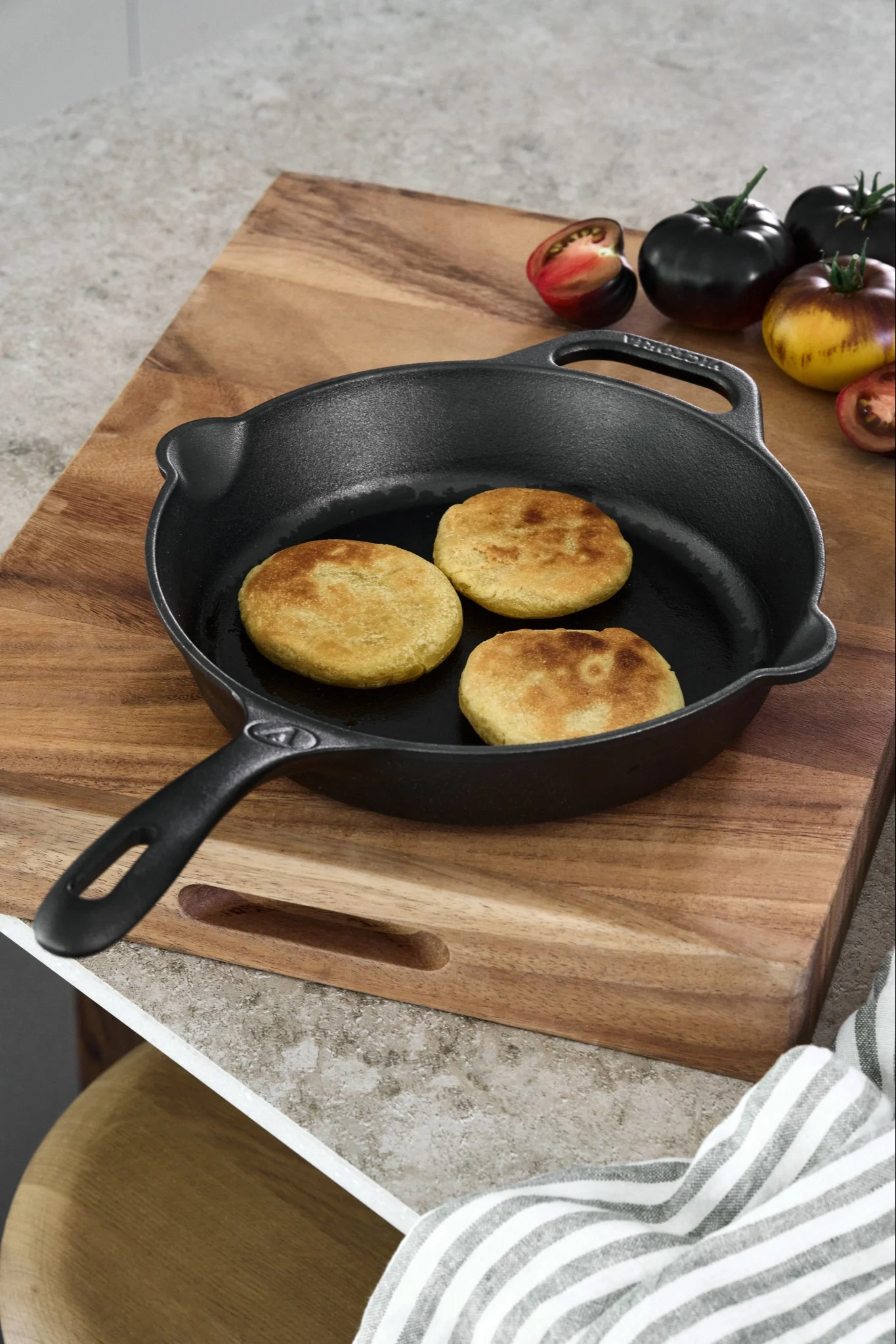A cast iron pan is placed on a wooden chopping board with a collection of tomatoes and fried arepas. 