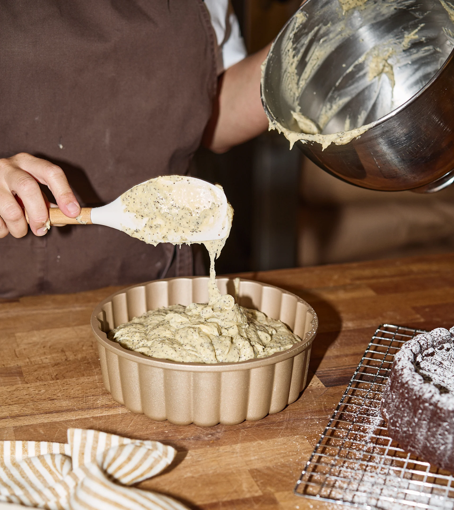 Person scooping poppy seed batter into a gold fluted baking pan. A chocolate cake with powdered sugar is in the background.