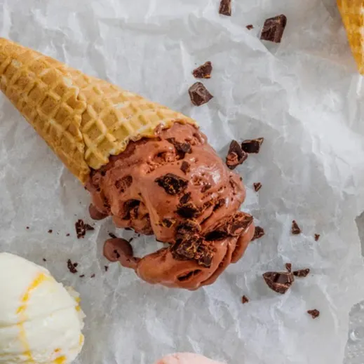 A melted chocolate ice cream cone with chocolate chunks on white paper, with a scoop of vanilla ice cream in the foreground.