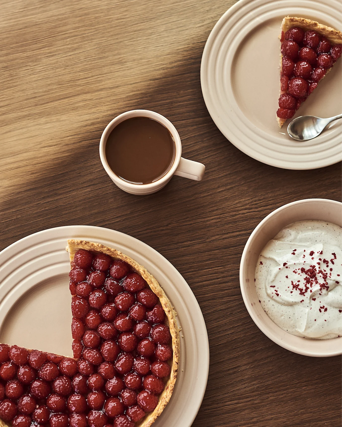 Berry tart with a slice removed, a slice on a plate, hot chocolate, and a bowl of cream on a wooden table.