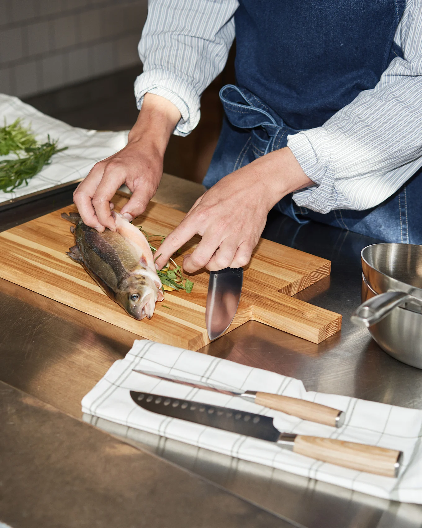 Hands preparing a whole fish on a wooden cutting board with a knife, next to other knives and a metal pot.