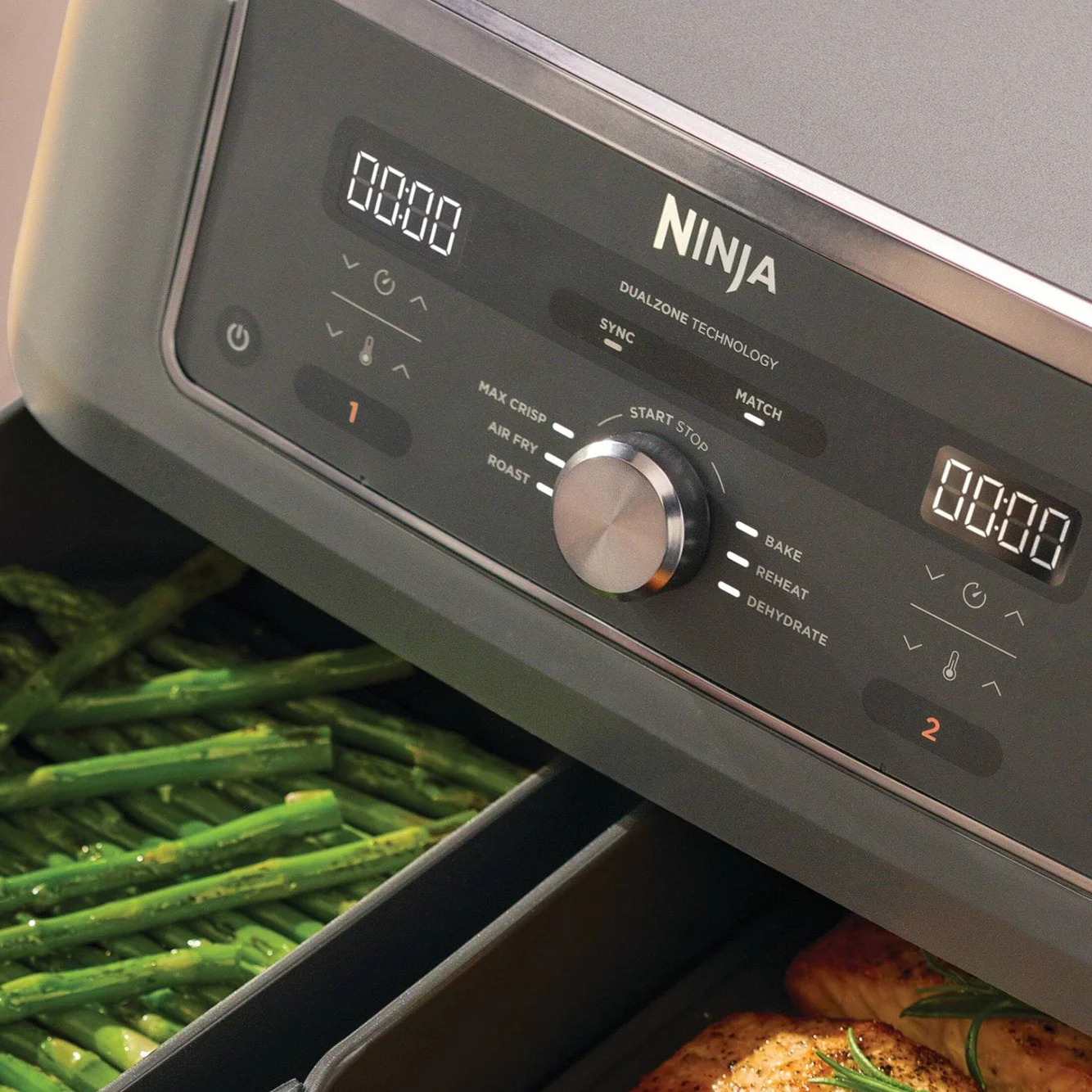 A Ninja air fryer's control panel with two digital displays and a knob, showing green asparagus in one drawer and chicken in another.