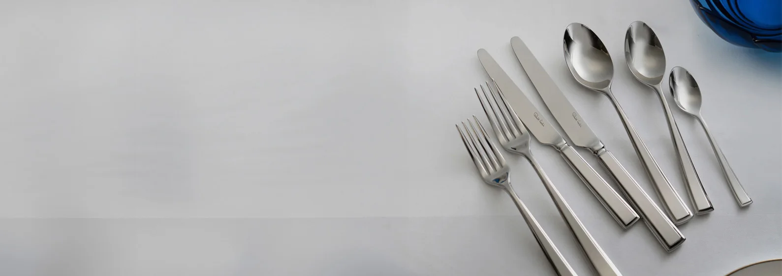 Silver cutlery set with forks, knives, and spoons on a white tablecloth, a blue glass bowl partly visible.