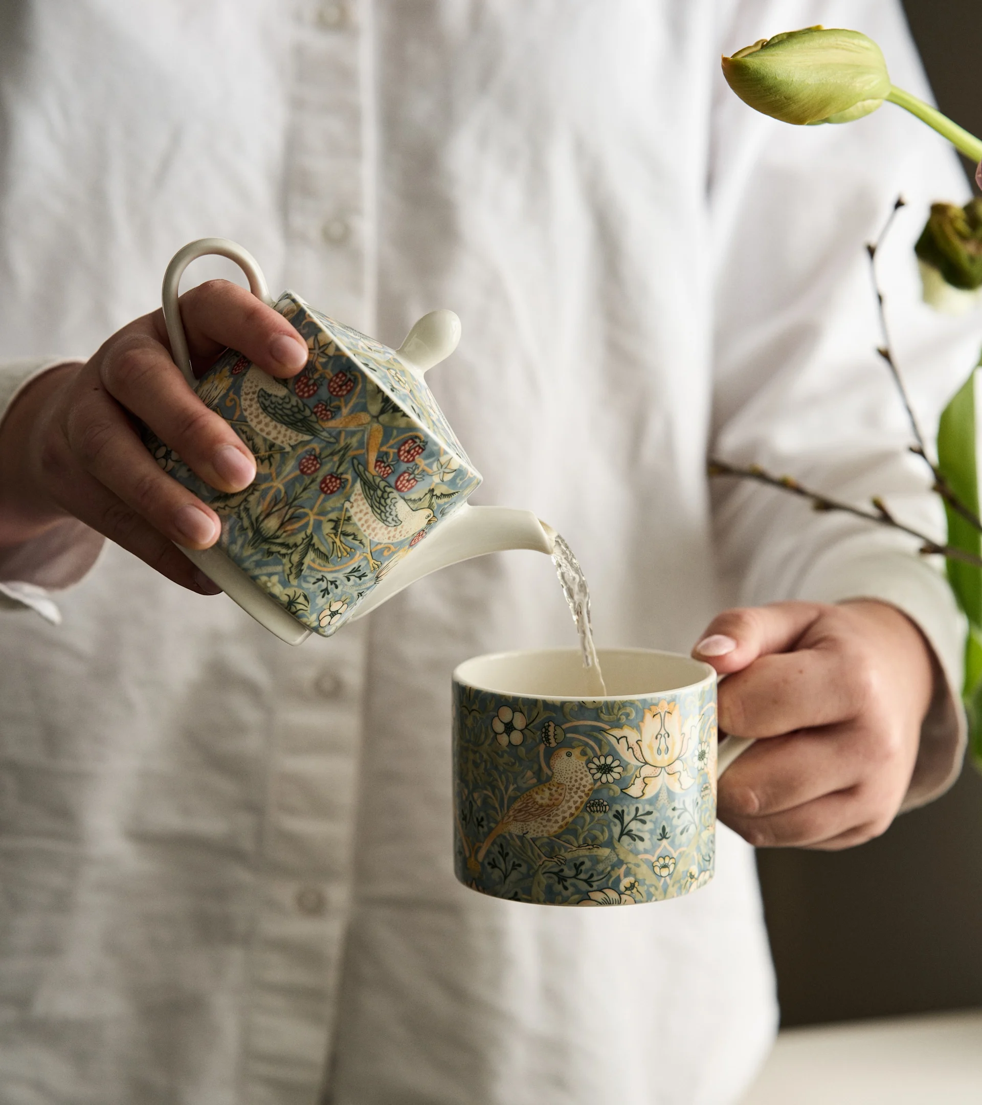 Person pouring water from a blue-green patterned teapot with birds and berries into a matching mug.