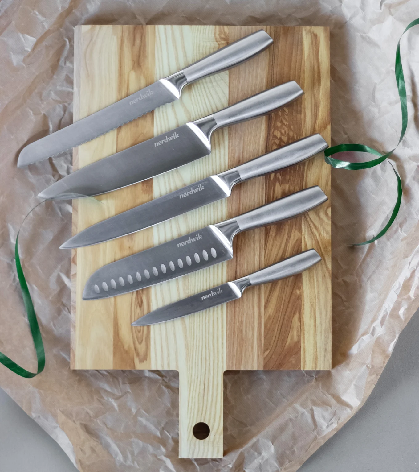 A set of five Nordwik stainless steel kitchen knives, varying in size, arranged on a light wooden cutting board with a green ribbon.
