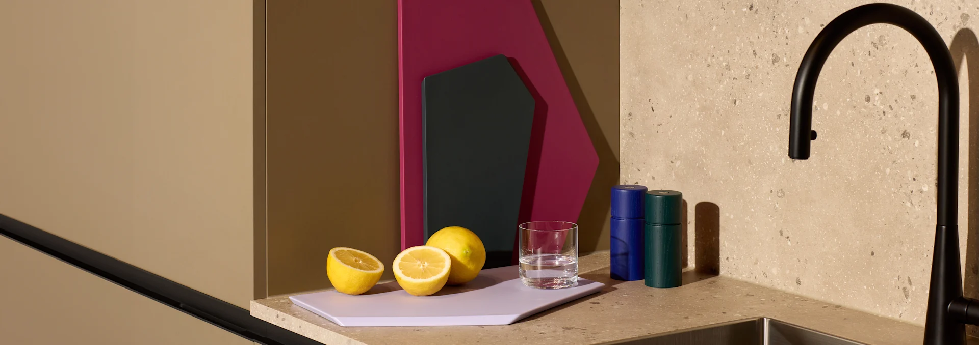 Lemons, water glass, and blue and green grinders on a purple cutting board on a speckled kitchen counter next to a black faucet.