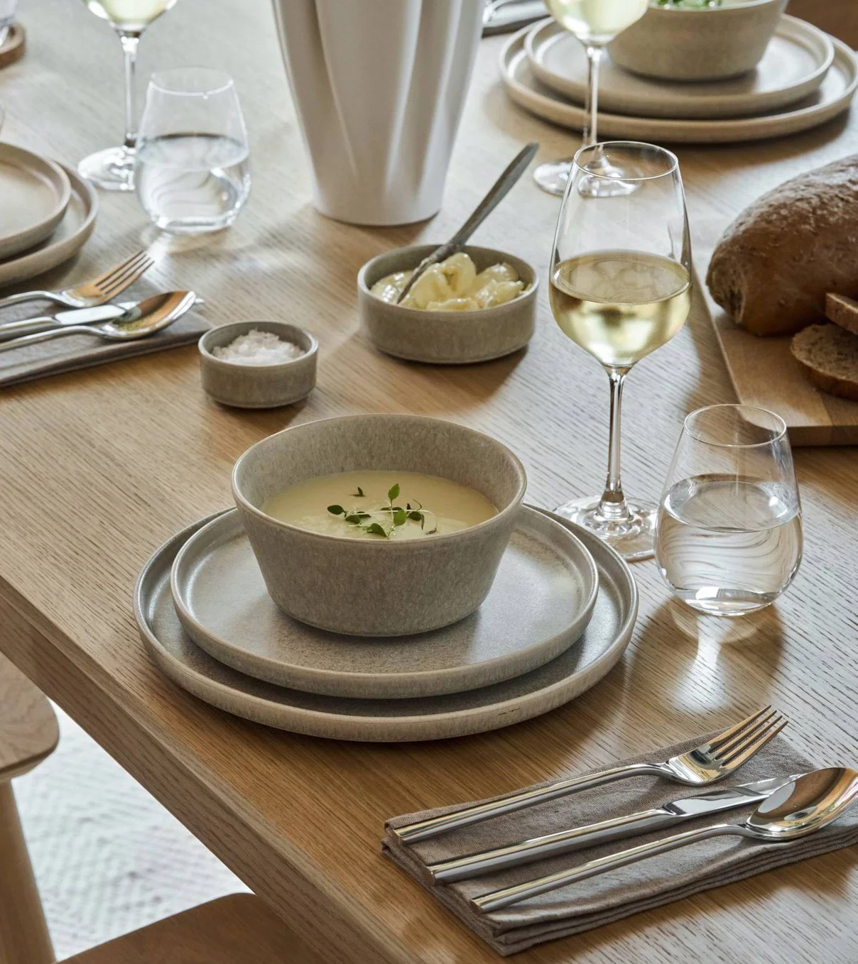 A dining table set with light grey stoneware, soup, bread, wine, water, and cutlery.
