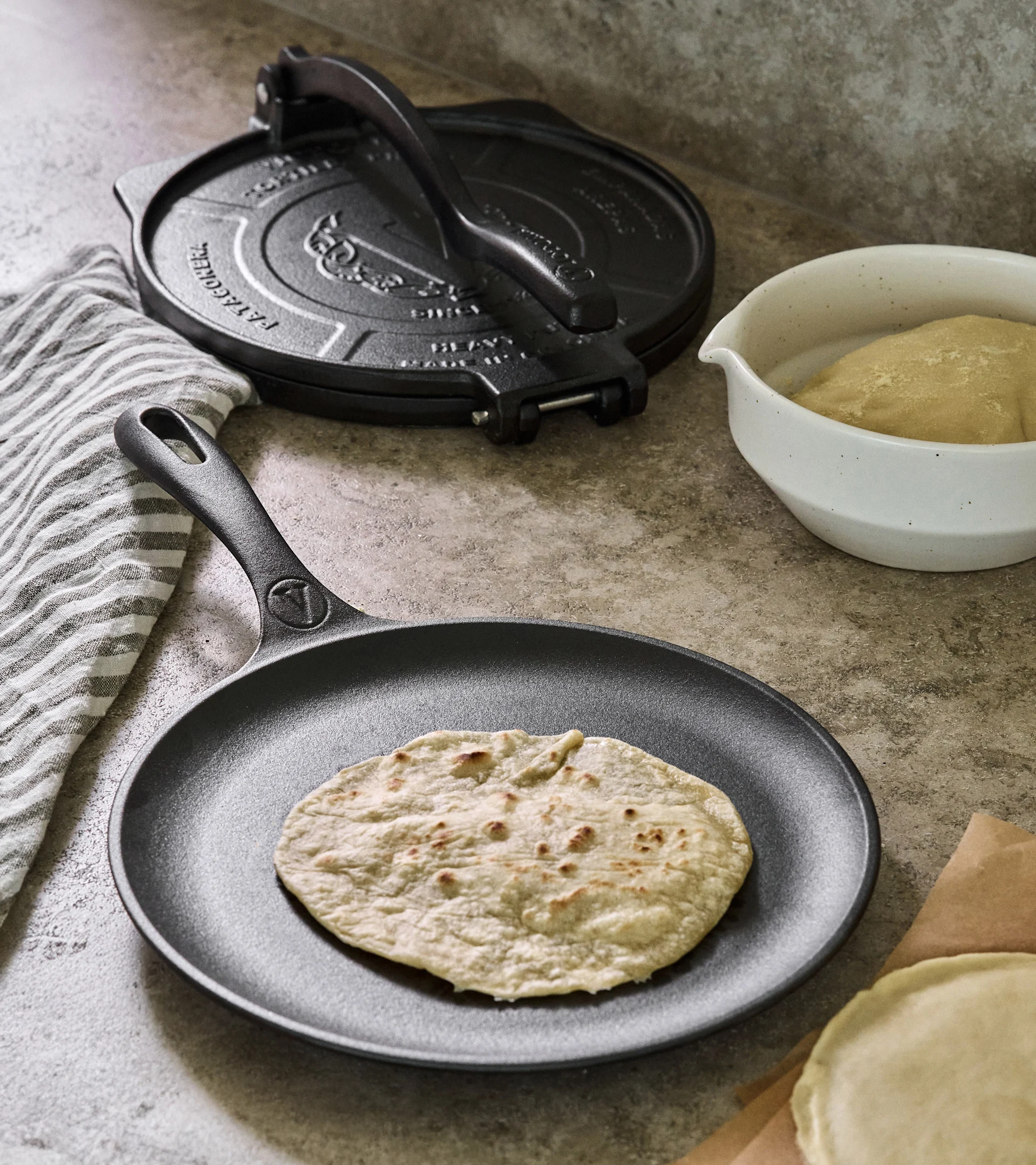 Cast iron tortilla press and skillet with cooked tortilla, dough, and uncooked tortillas on a counter.