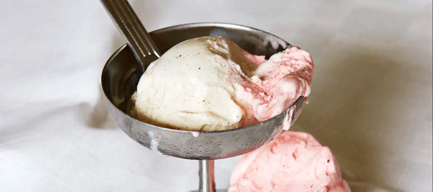 Two scoops of ice cream, vanilla bean and strawberry, in a metal sundae dish with a spoon.