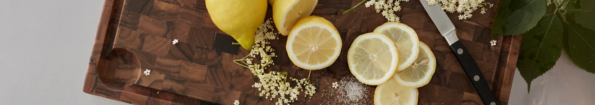 Lemons, elderflower, and sugar on a wooden cutting board with a knife.