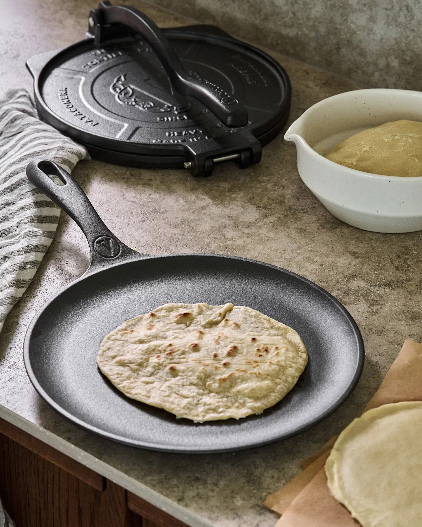 A cooked tortilla rests on a black cast iron griddle, with a tortilla press, dough bowl, and uncooked tortilla in the background.