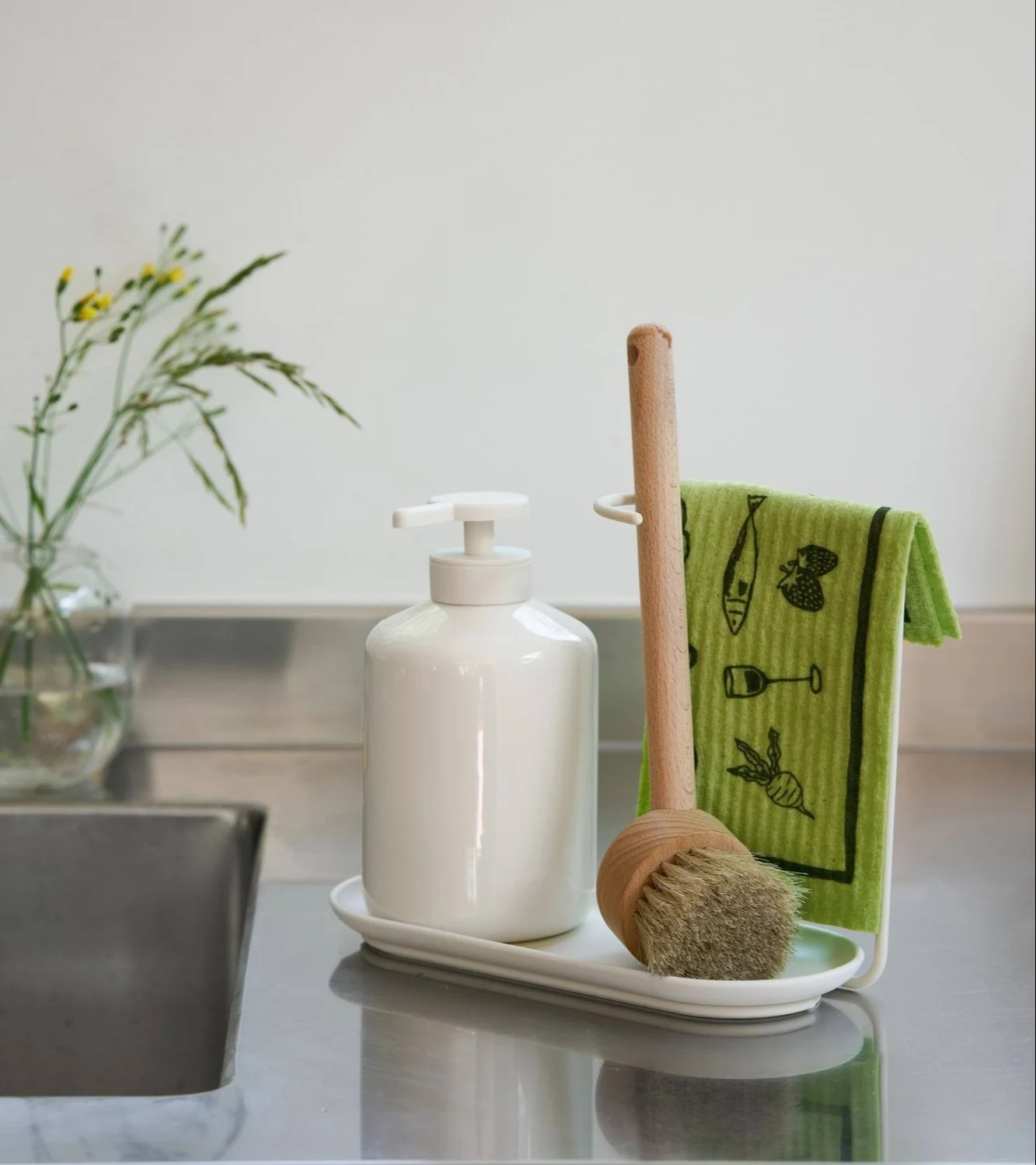 A white ceramic soap dispenser, a wooden dish brush, and a green dishcloth with illustrations on a tray next to a sink.