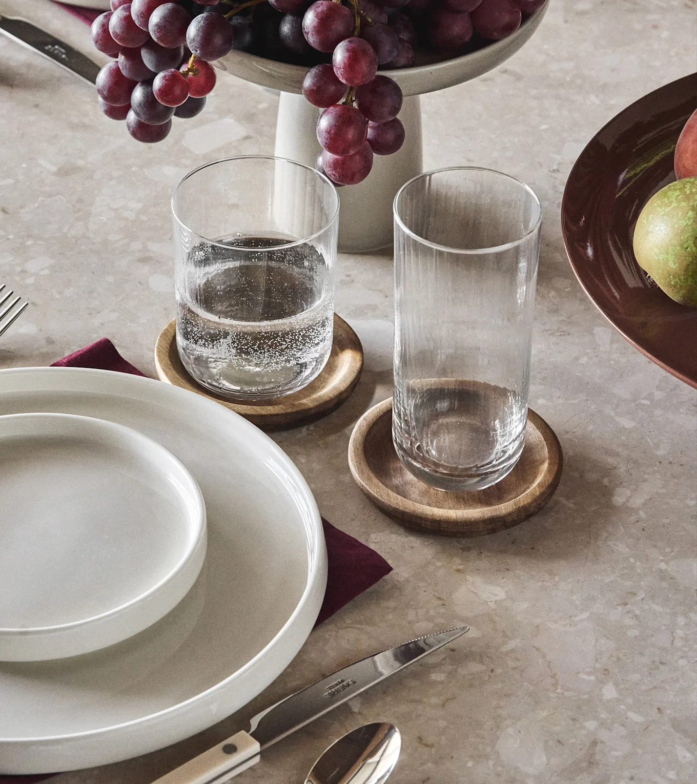 Dining table with white plates, two clear glasses on wooden coasters, red grapes, and a piece of fruit.