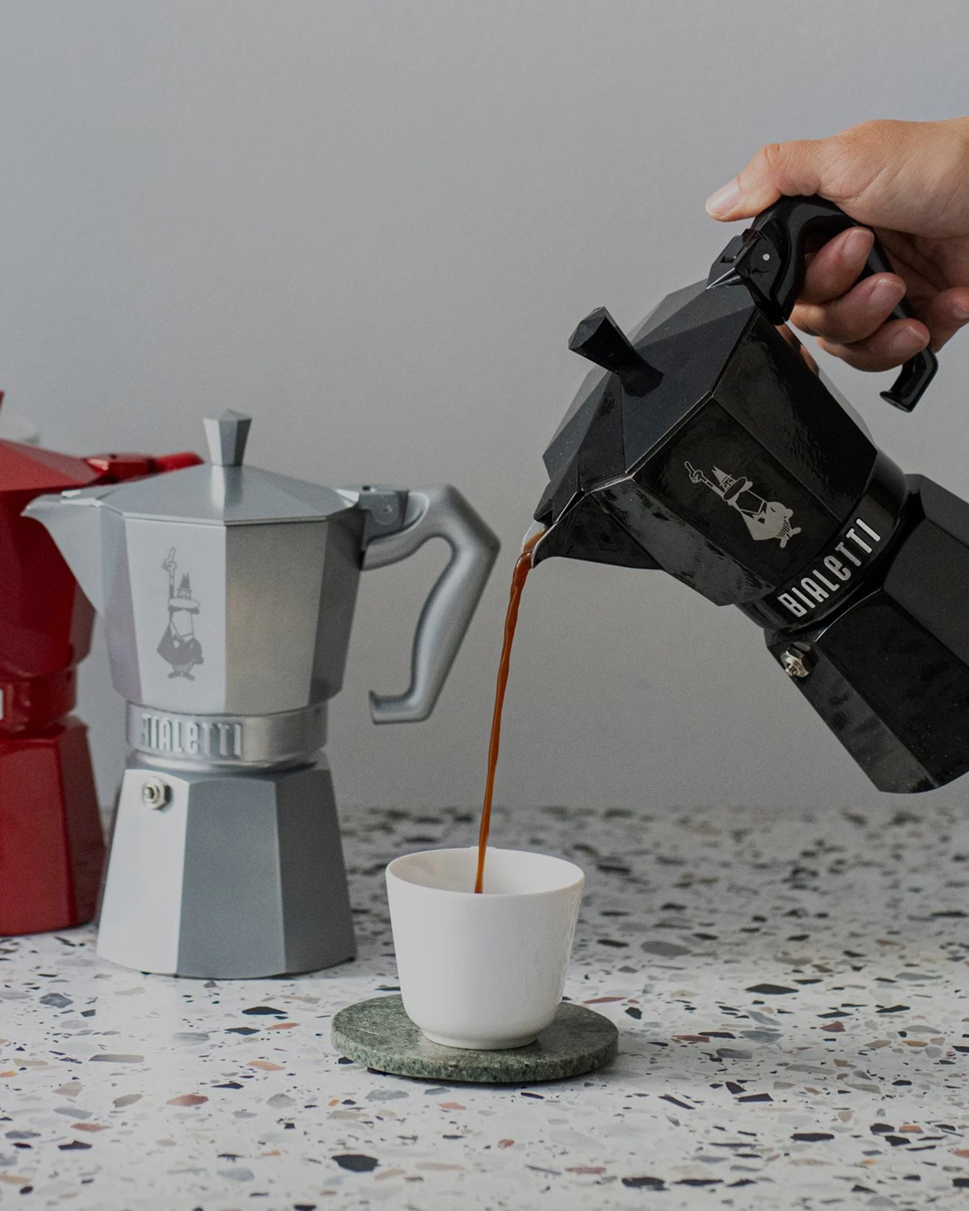 Hand pouring coffee from a black moka pot into a white cup, with silver and red moka pots in background on a speckled counter.